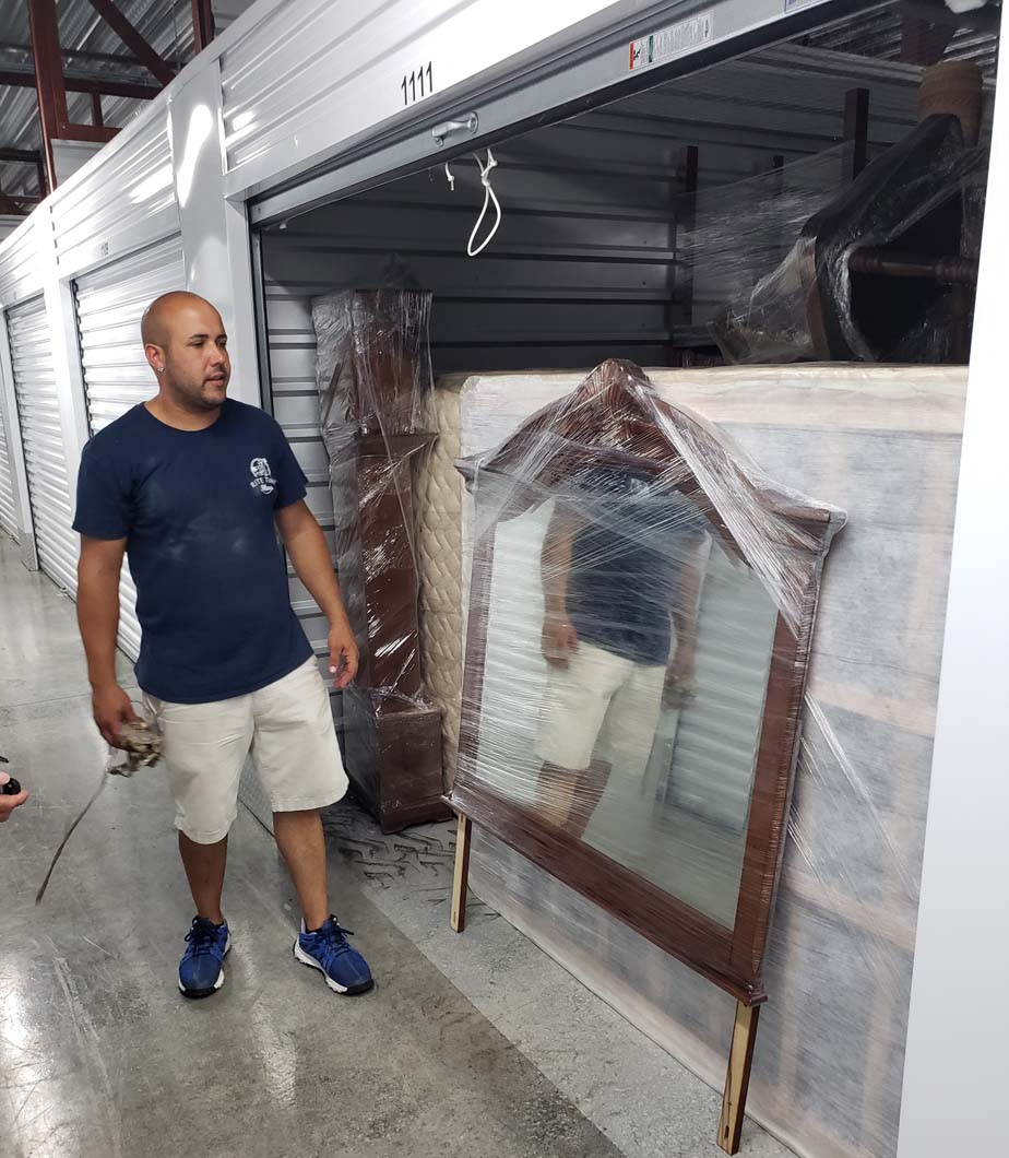 Man in storage unit, standing near wrapped furniture, including a mirror.