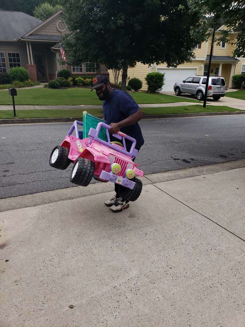 Man carrying a pink toy Jeep on a driveway. Houses and a parked car are in the background.