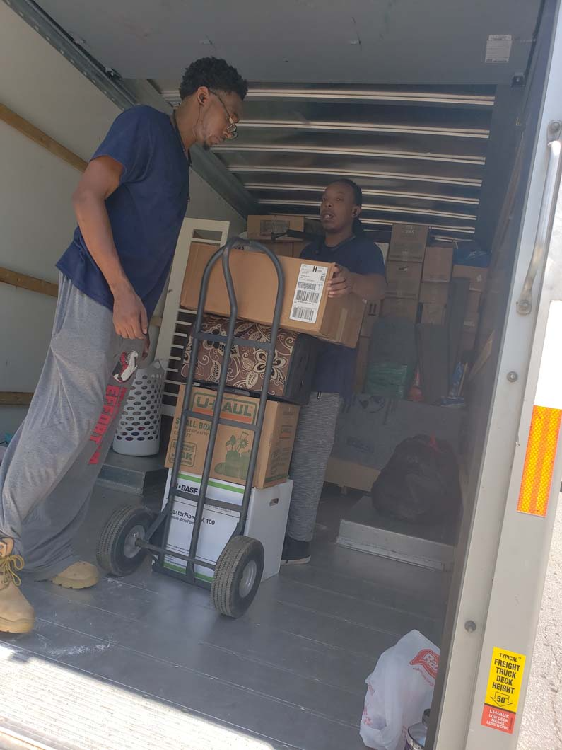 Two people loading boxes into a moving truck. One uses a dolly. Bright sunlight.