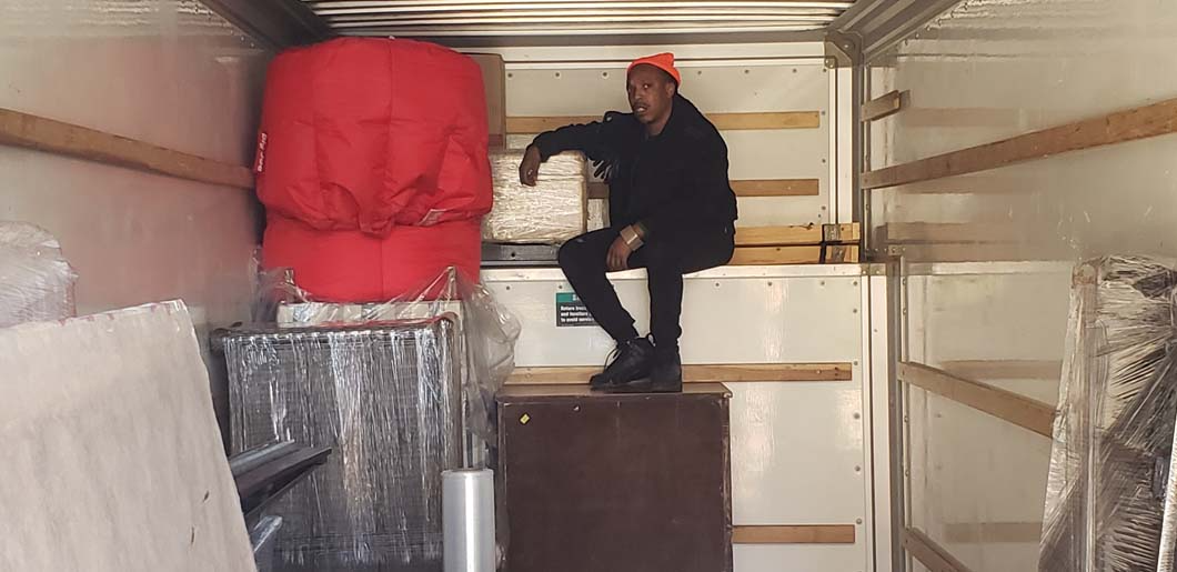 Person in orange hat sits in a moving truck, next to a red bag. Interior shot.