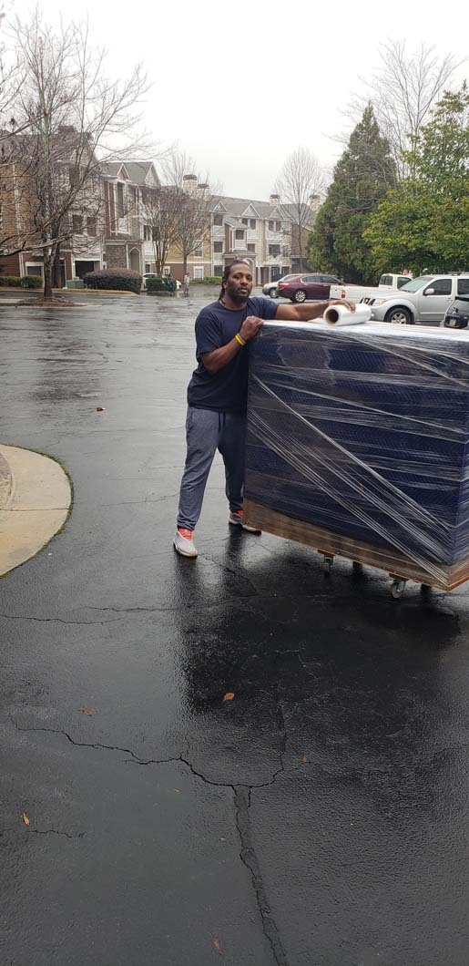 Person pushing a large, wrapped blue object on a dolly across a wet asphalt surface. Buildings in the background.
