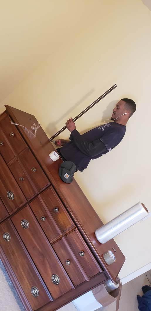 A person leans against a dresser holding a long, thin object. The wall is light yellow.