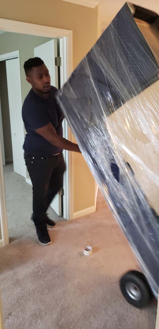 Person moving a large, wrapped item on a dolly through a doorway in a carpeted room.