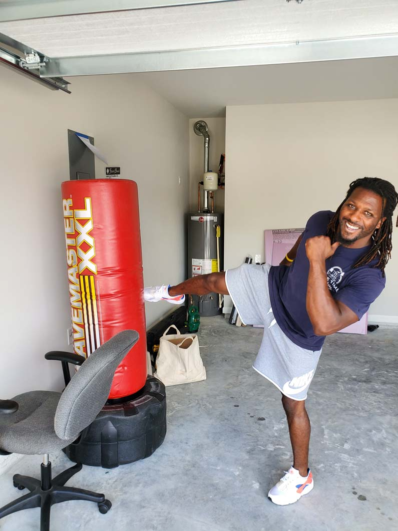 Man kicks a punching bag in a garage. He is wearing a dark shirt and light shorts with white sneakers.