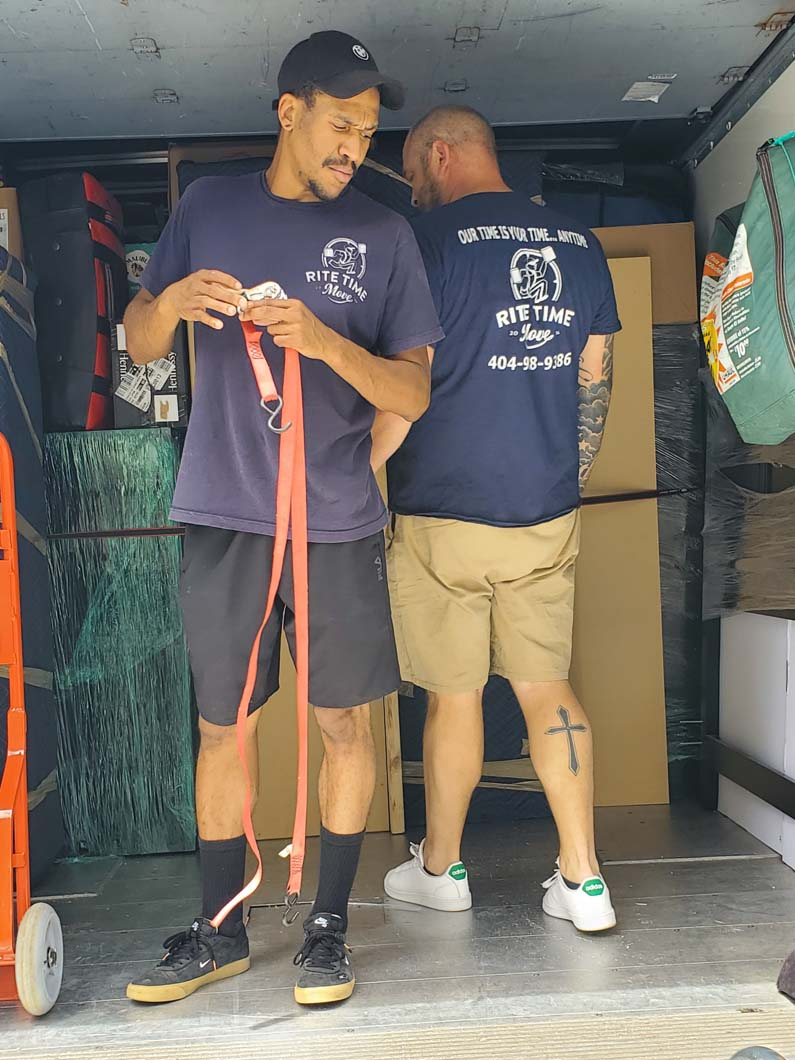 Two movers inside a truck, one holding straps, other near boxes. Both wear navy shirts with logo, shorts, sneakers.