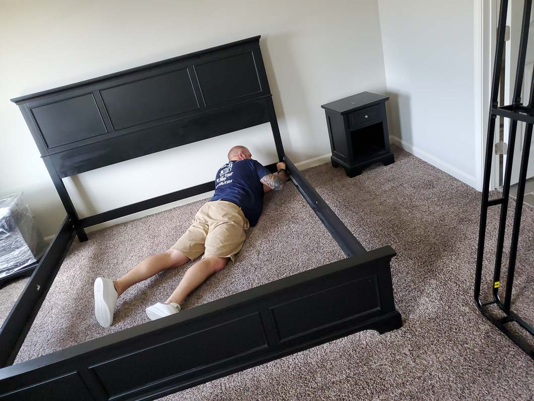 Man lying inside a black bed frame. Headboard upright, nightstand beside it. Beige shorts, white shoes.