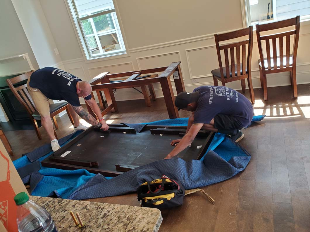 Two movers assembling a disassembled table on a blue moving blanket. Wooden chairs sit nearby.