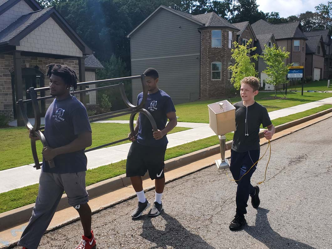 Three people carrying items down a residential street; one with a box, two with metal hoops.