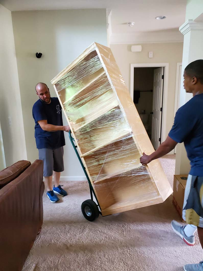 Two movers transport a large bookcase on a dolly through a house, on carpet.