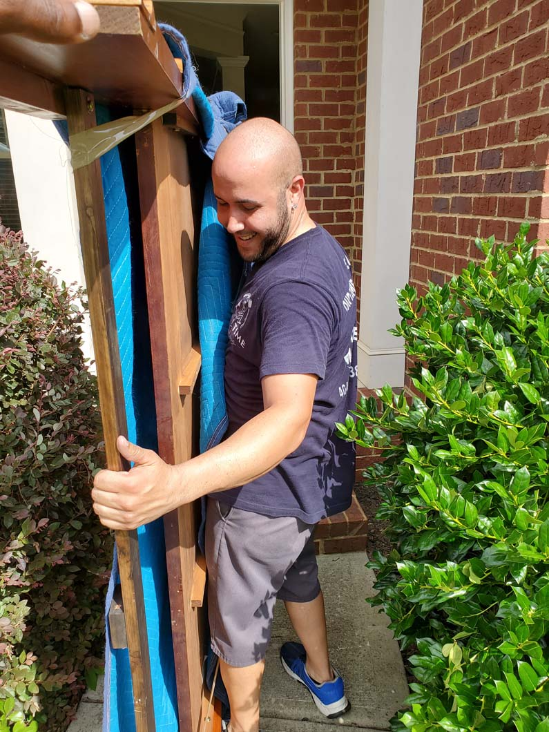 Man carrying a tall, vertical wood object wrapped with blue fabric, smiling outside a doorway.