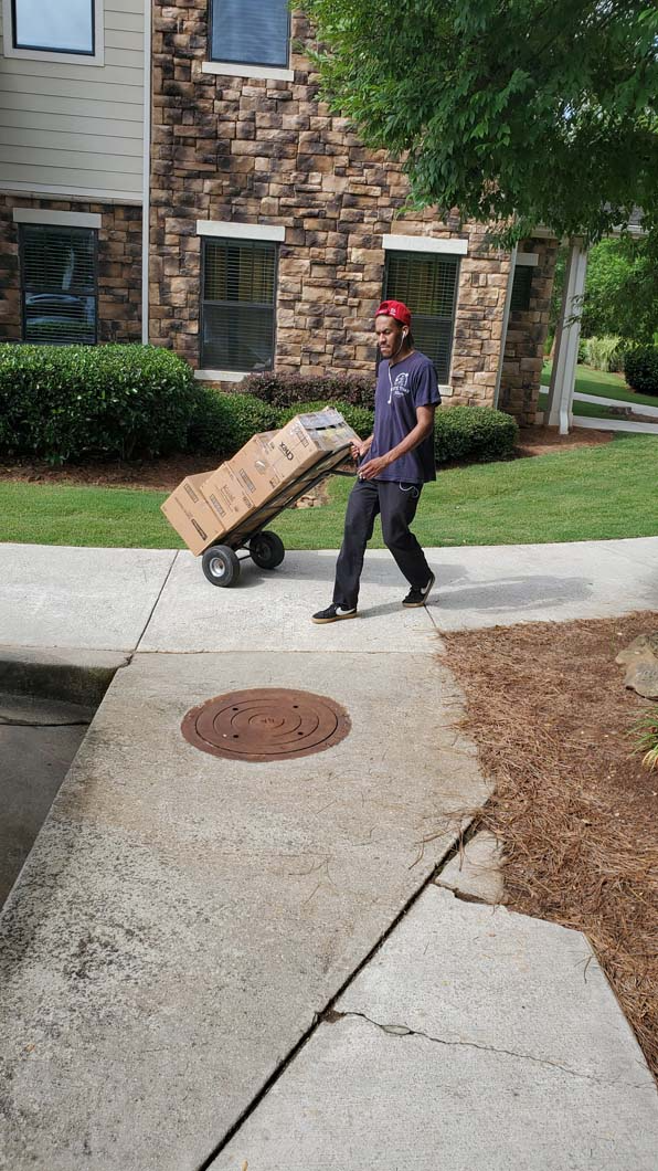 Person pulling a large cardboard box on a wheeled dolly on a sidewalk outside a building.
