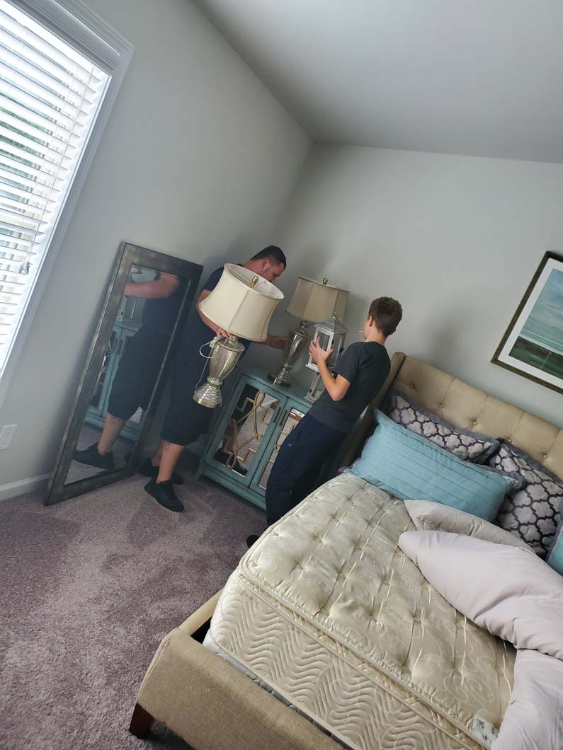 Two people moving furniture in a bedroom. One holds a lamp, the other a mirrored chest. A large mirror and bed are also present.