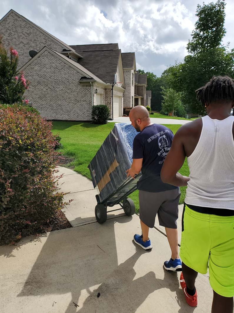 Two people moving a large box on a dolly up a driveway towards a house.