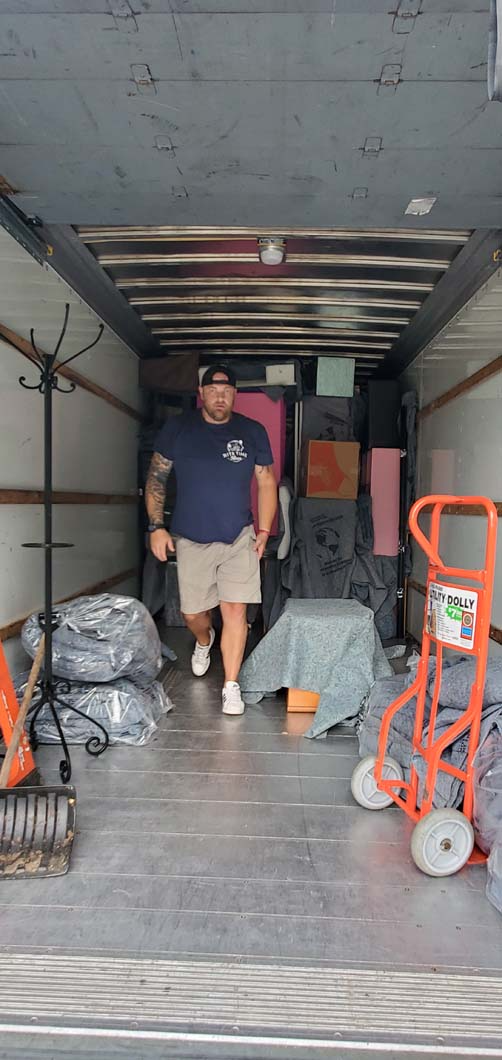 Man walking out of a moving truck filled with furniture.  An orange dolly and wrapped items are visible.