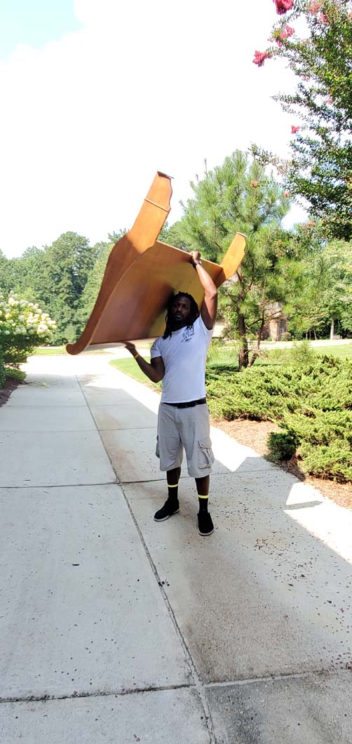 A person holds a large, brown, wooden object above their head on a sunny sidewalk.