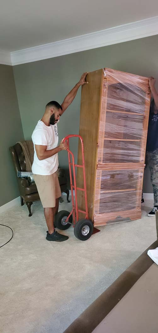 Two people moving a large, wrapped cabinet with a hand truck indoors.