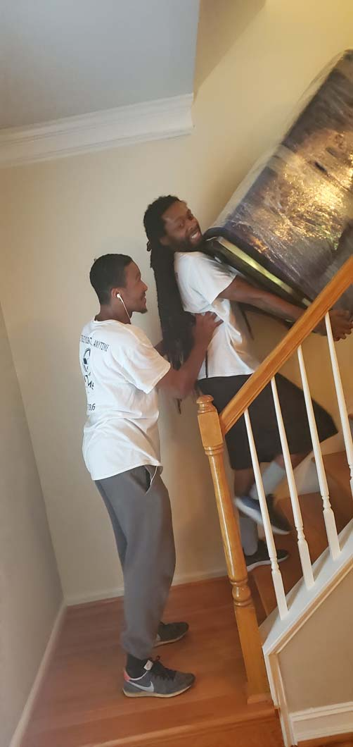 Two people carry a large, wrapped object up wooden stairs. One person is in front, the other behind, assisting with the lift.