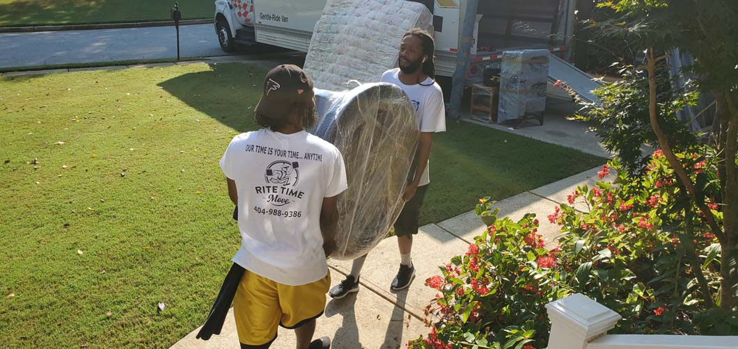 Two movers carrying a wrapped round table on a grassy lawn near a moving truck and bushes.