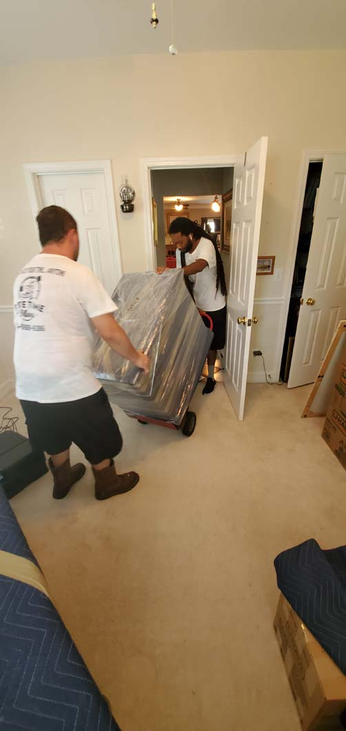 Two movers carry a wrapped piece of furniture through a doorway.