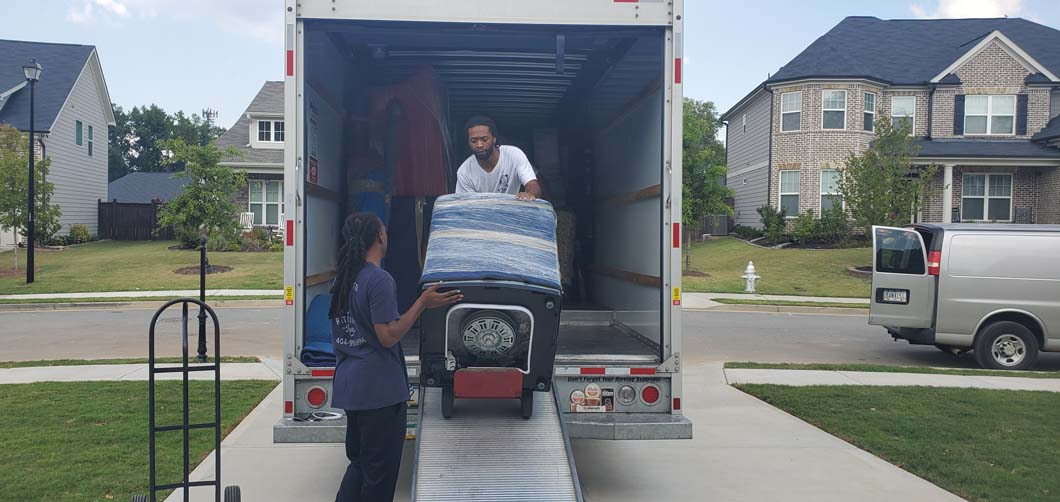 Movers loading a truck with a dolly on a driveway. Houses in the background. Sunny day.