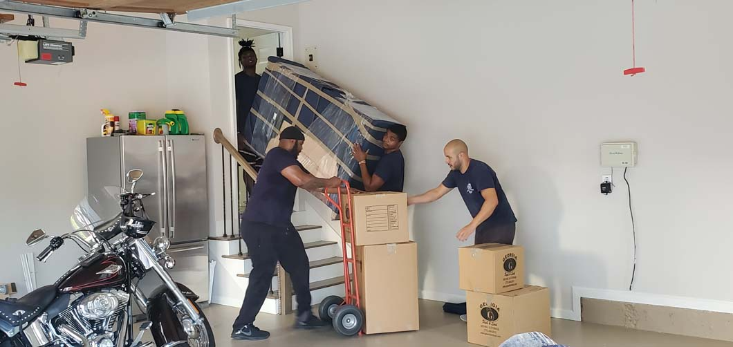 Three people moving furniture and boxes into a garage. One pushes a dolly. Others help with a mattress and boxes. Motorcycle parked.