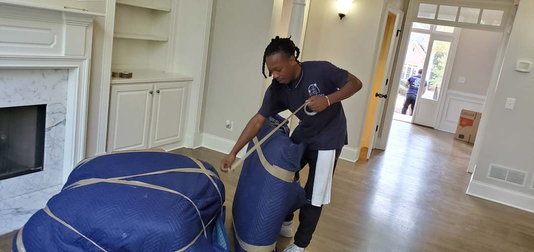 Movers wrapping furniture in a new home. One person holds a strap to secure blue-covered items.