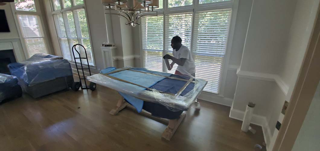 Person wrapping a rectangular table in plastic, in a room with large windows.