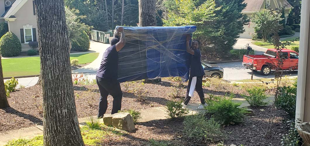 Two people carrying a large, wrapped item through a yard. A red truck is parked nearby.