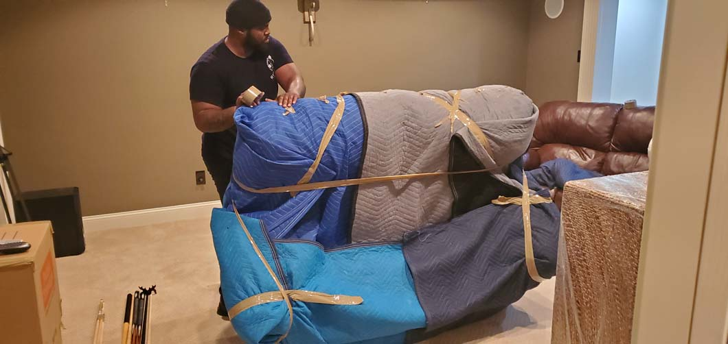 A person wrapping furniture with blue and gray covers and securing it with straps. Indoors.