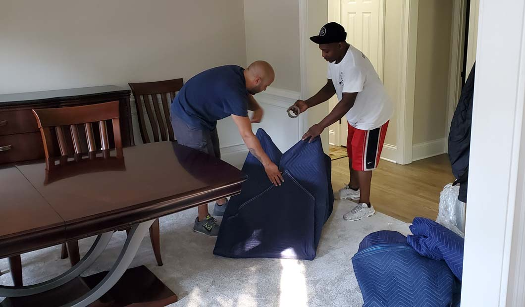 Two people wrapping furniture in a room with dining table. One is wearing a white shirt and the other a blue shirt.