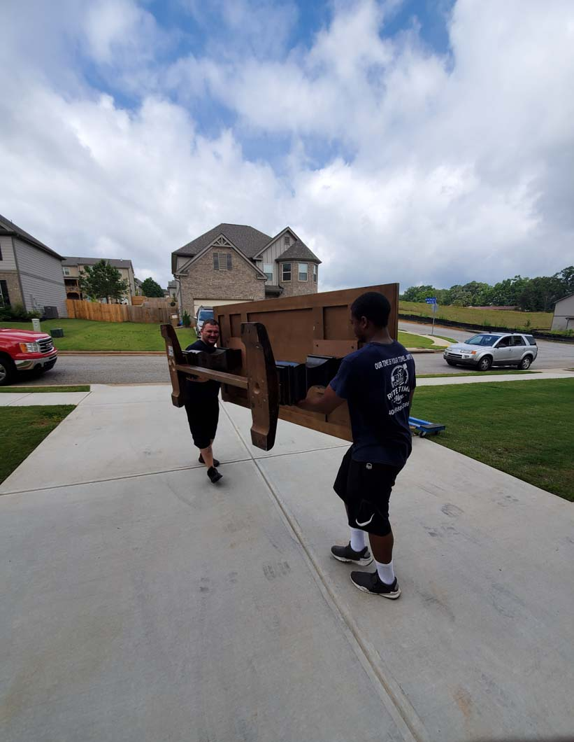 Two people carrying a wooden table on a driveway in front of a house. Cloudy sky.