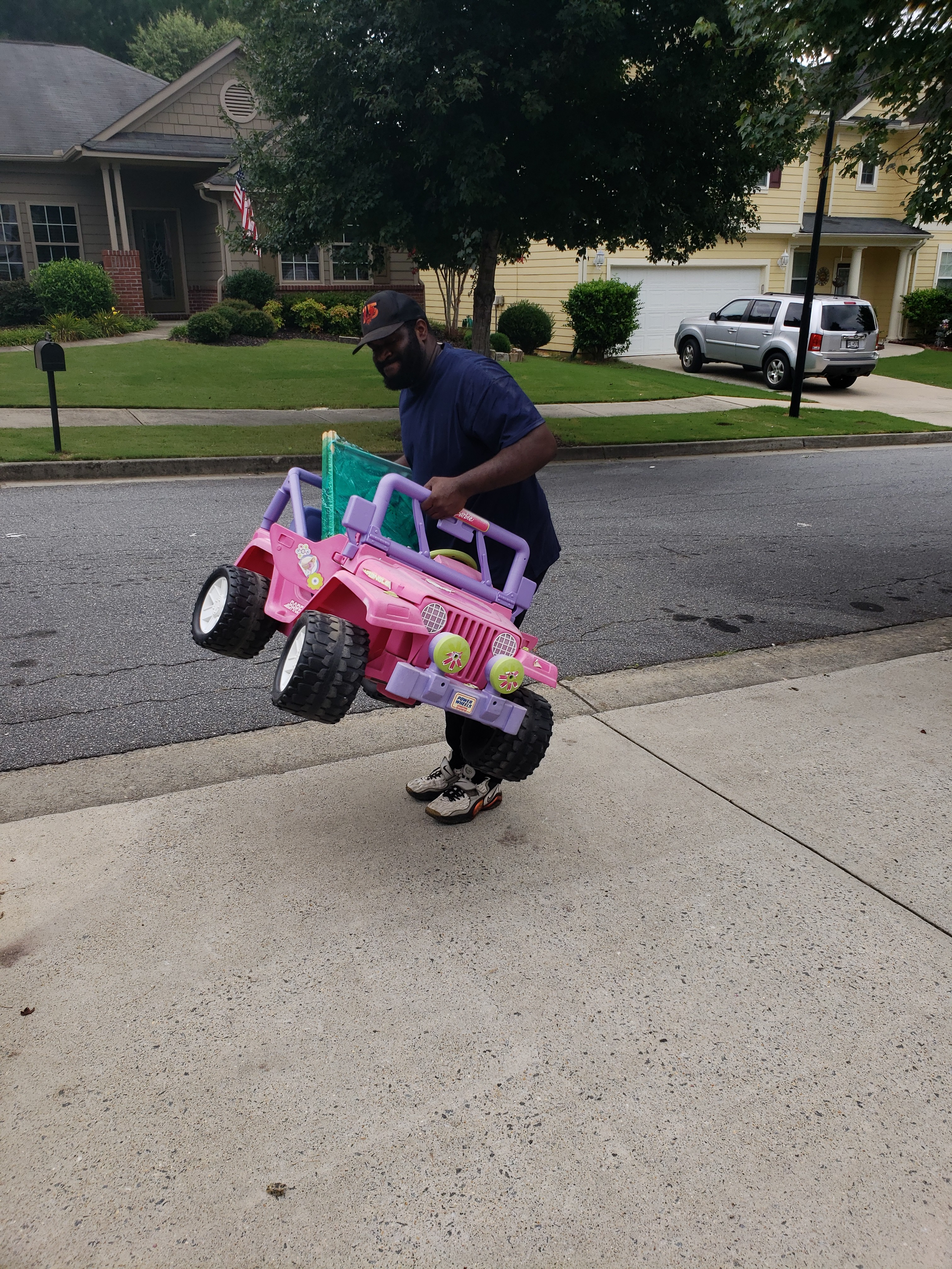 Man carrying a pink toy jeep on a sidewalk in front of houses.