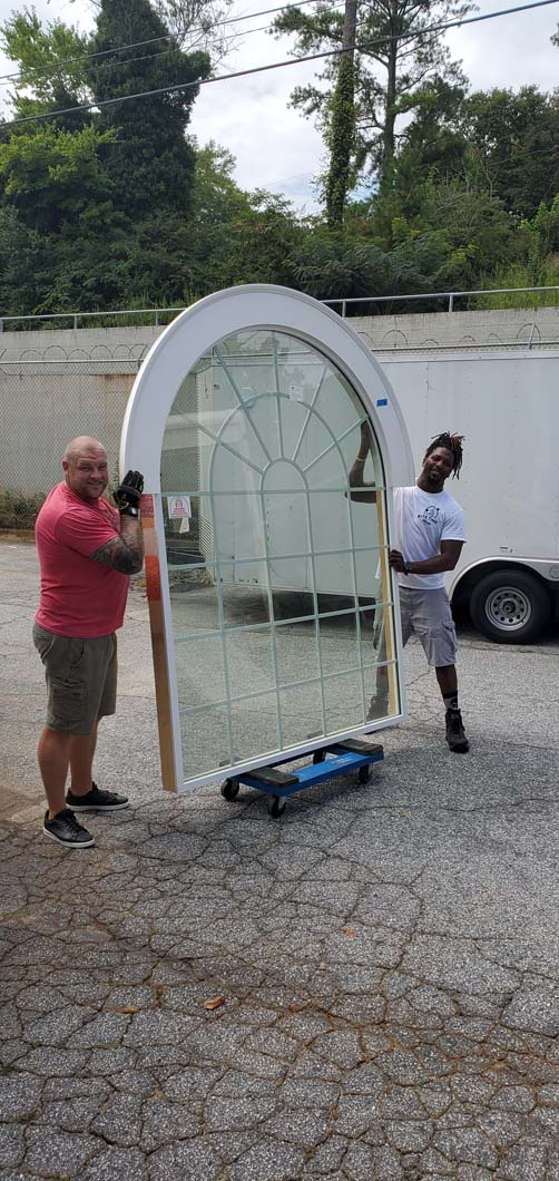 Two people hold a large, arched window with a dolly on a gravel surface.