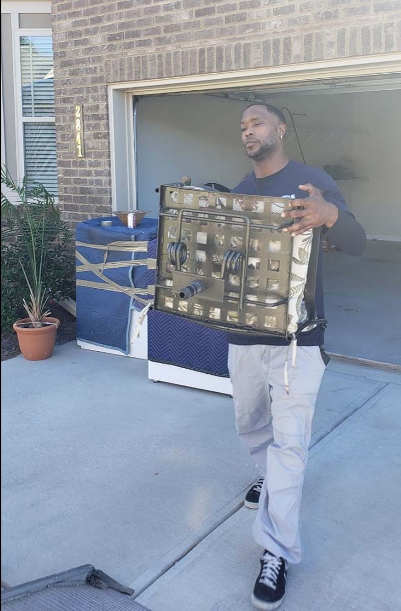 Man carrying a large, dark metal object outside a garage, a blue and white object in the background.
