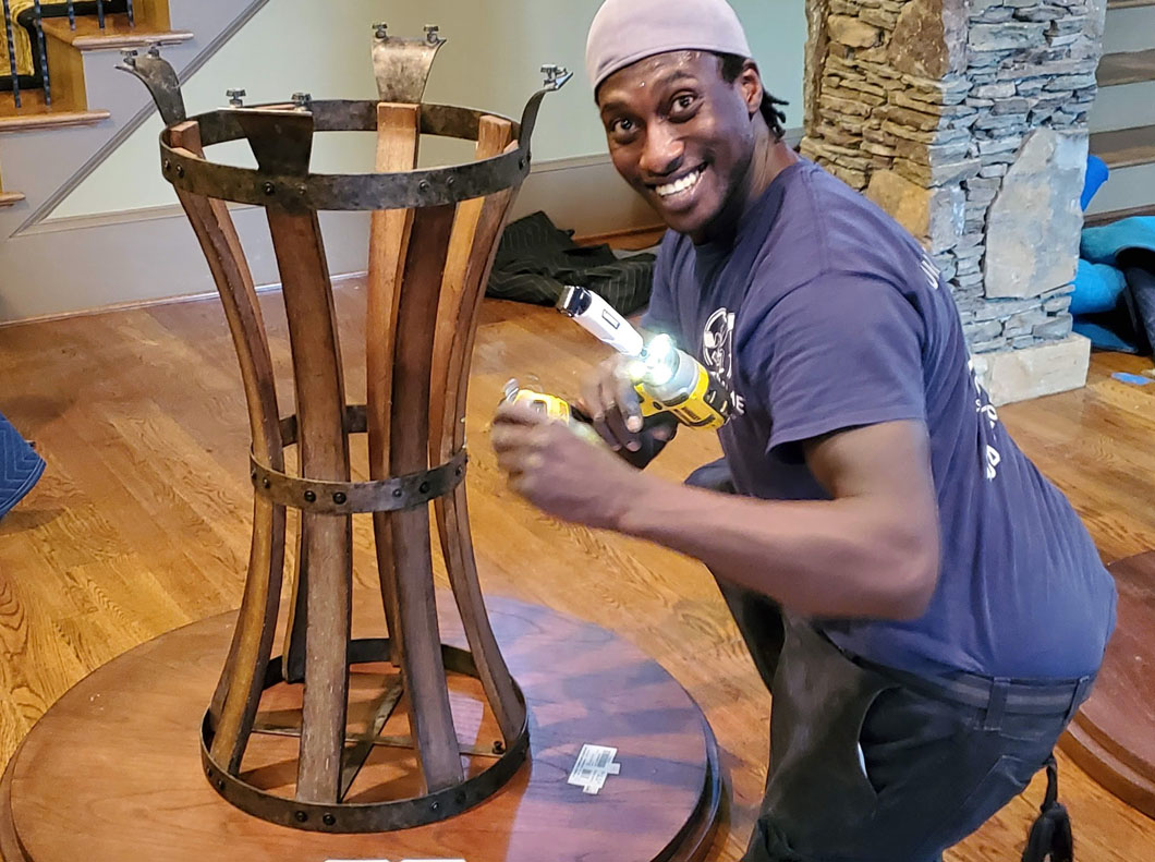 Man assembling table base with a drill, smiling. Wooden barrel-shaped base on round table, interior setting.