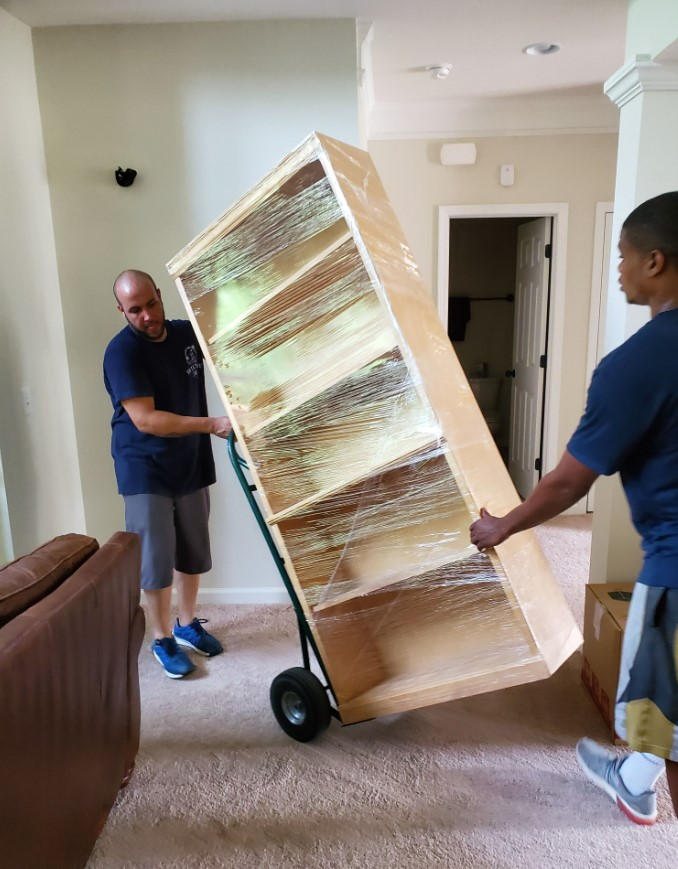 Two people moving a large, wrapped bookcase on a hand truck through a room with carpet flooring.