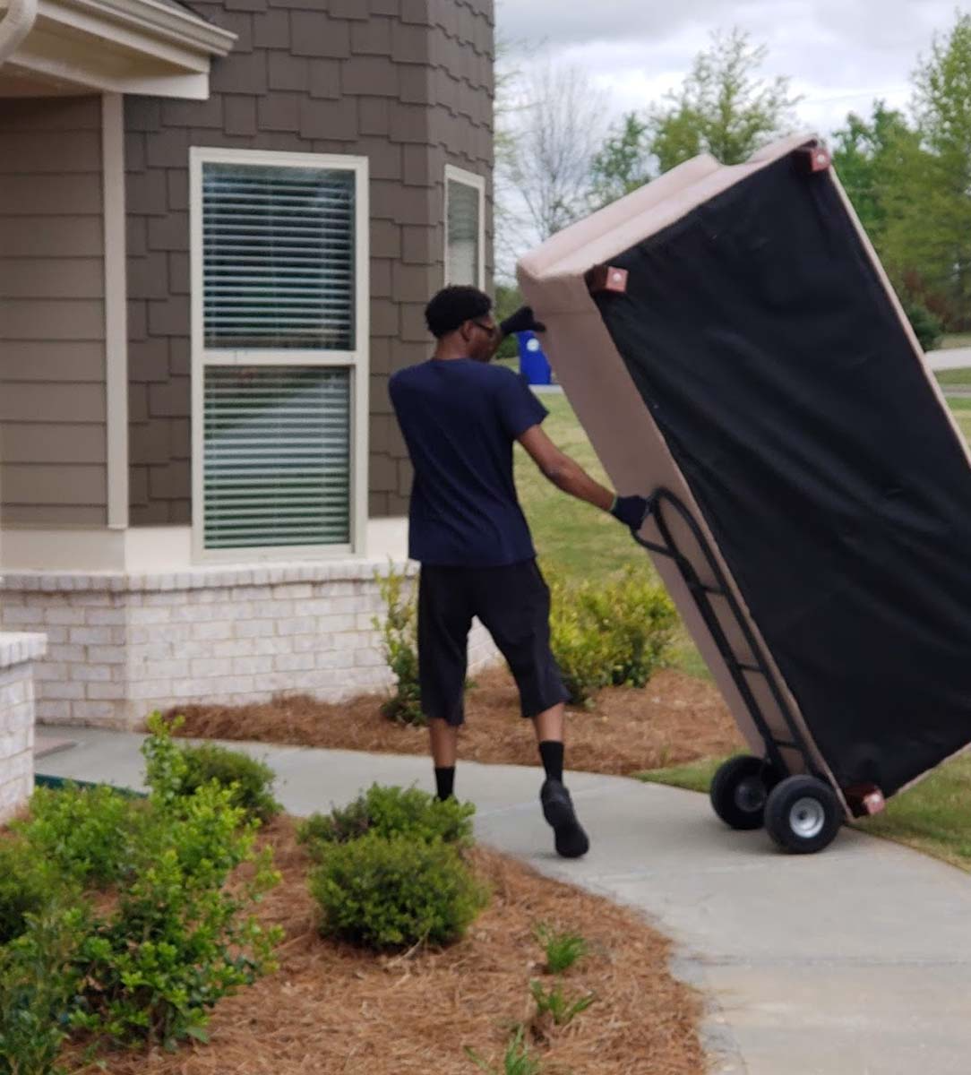 Man using a dolly to move a large, covered object on a sidewalk near a house.