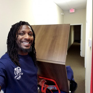 Man with dreadlocks smiling, holding furniture on a hand truck in hallway.