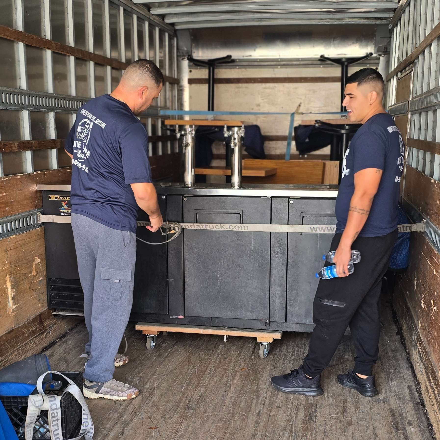 Two movers securing a black refrigerator on wheels inside a truck.