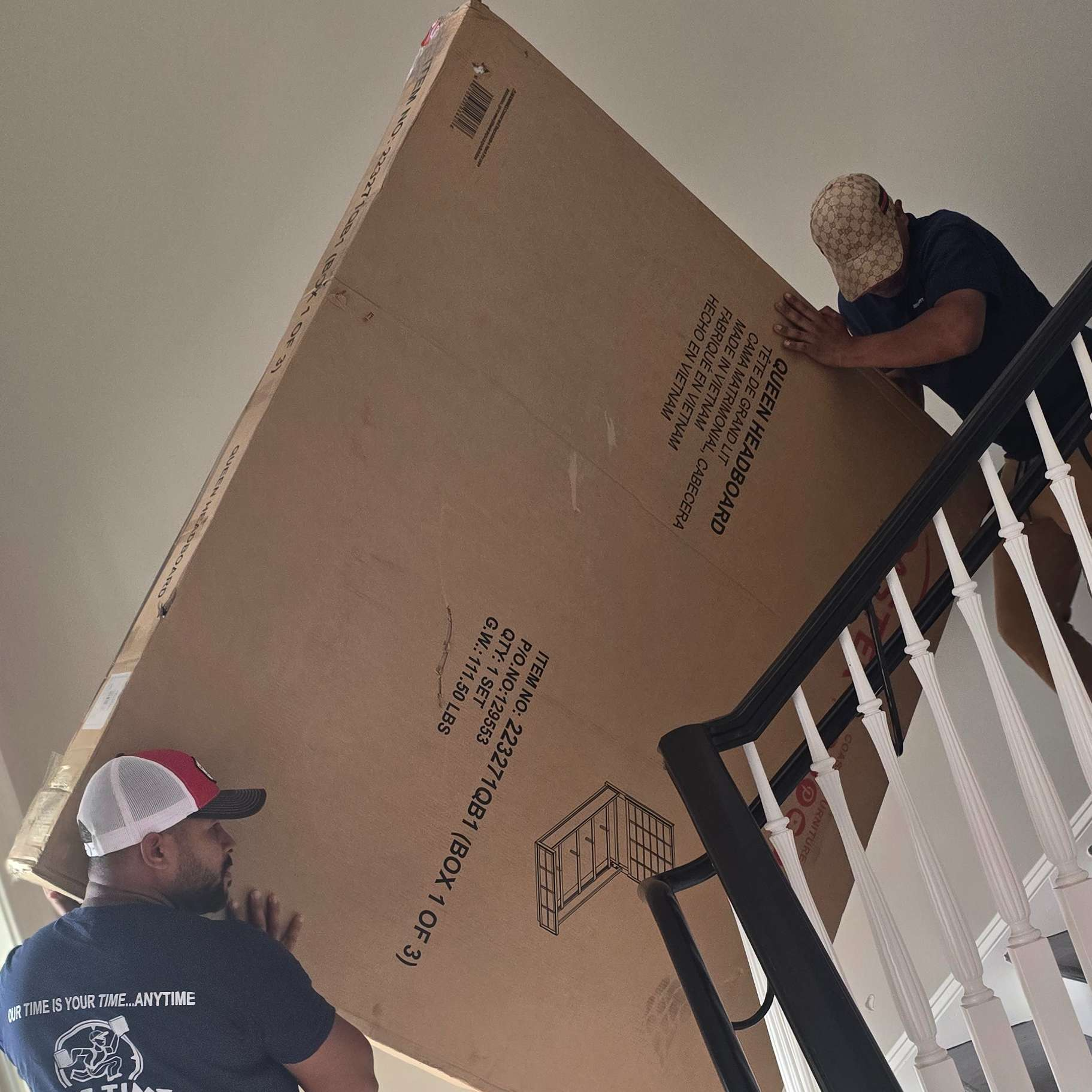 Two men carrying a large, rectangular cardboard box up a staircase.