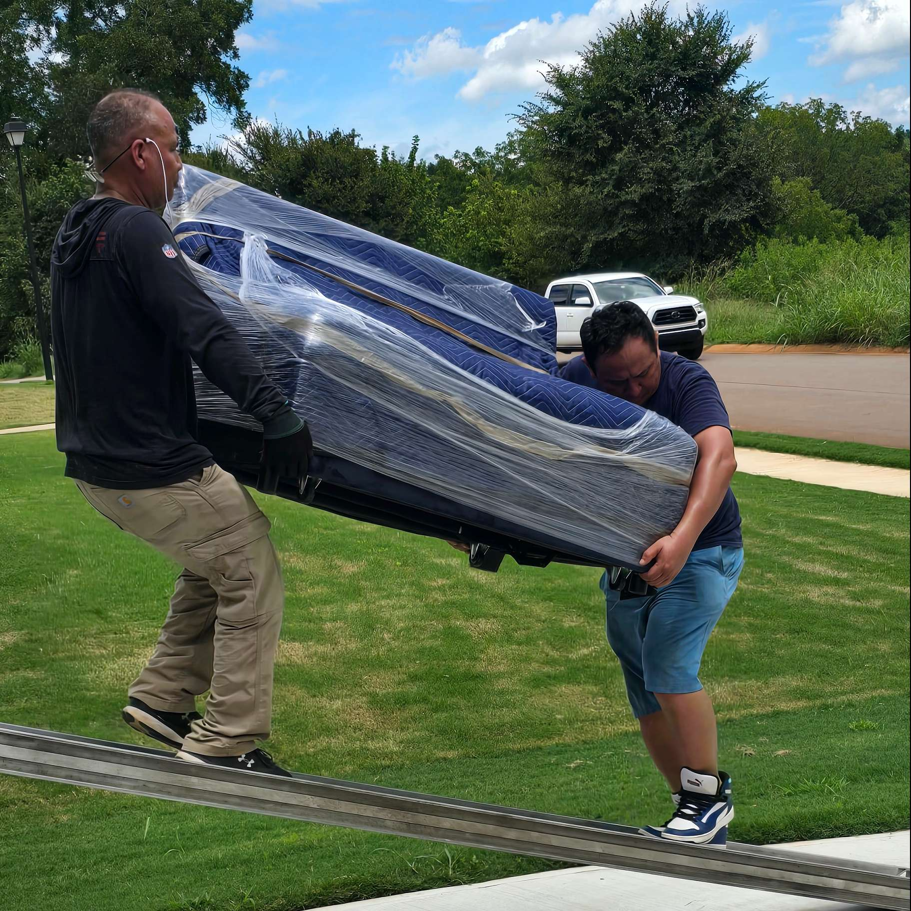 Two people carrying a large, blue-wrapped couch on a grassy lawn.