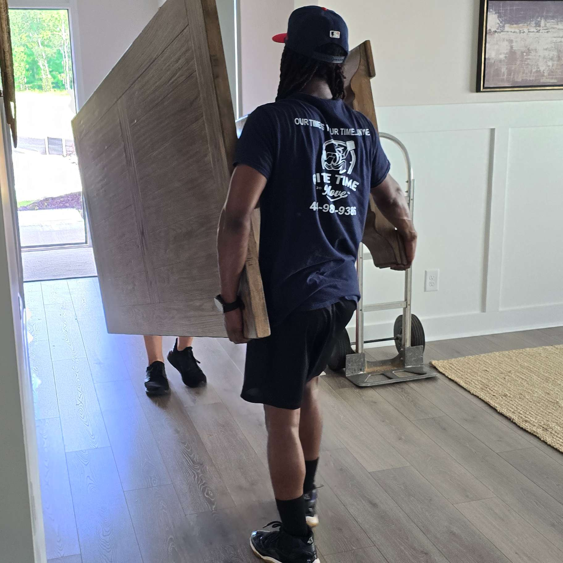 Two people carrying large wooden panels through a doorway. One wears a blue t-shirt and black shorts.