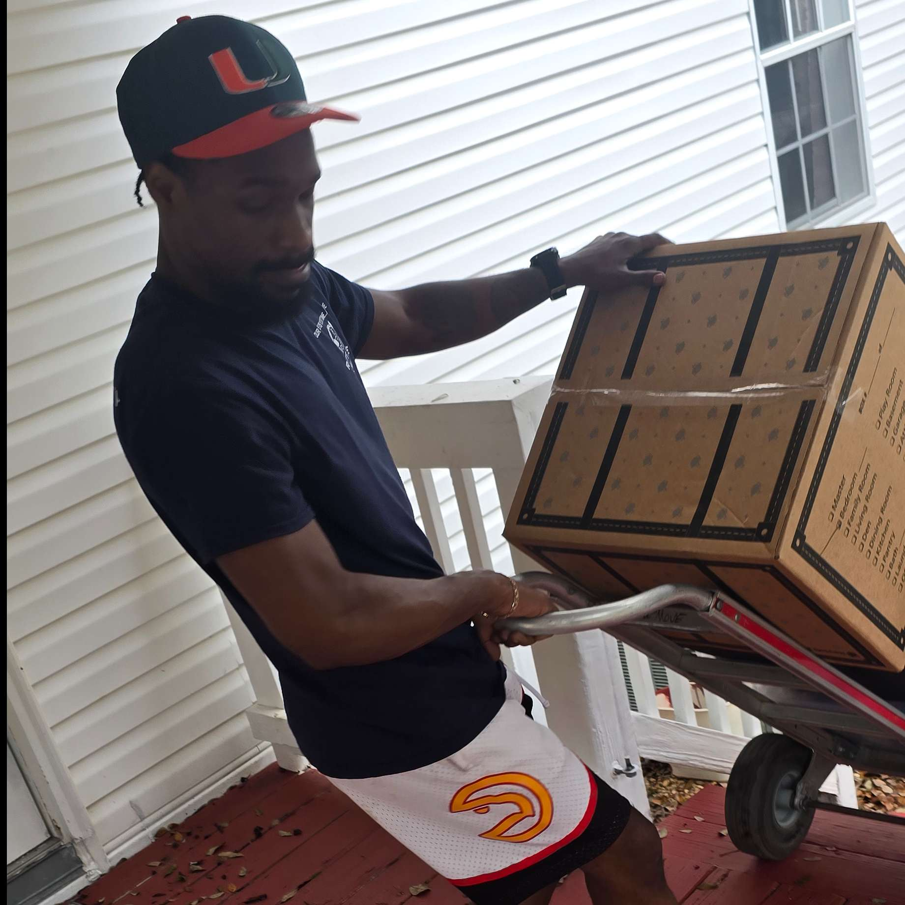 Man in black shirt and Hawks shorts, pulling box on a dolly up porch steps.