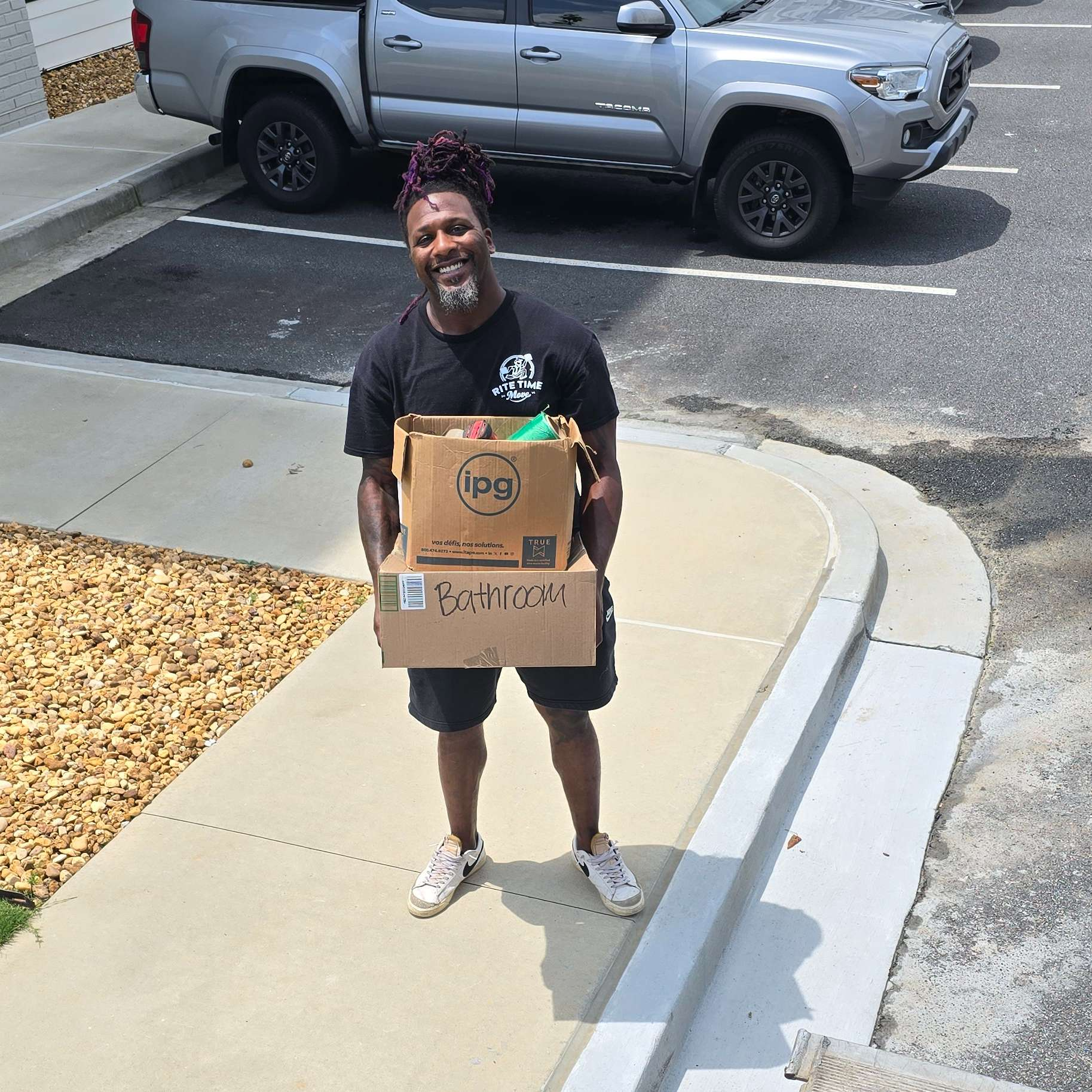 Man holding two boxes, smiling outside near a truck. He is wearing a black shirt, shorts, and sneakers.