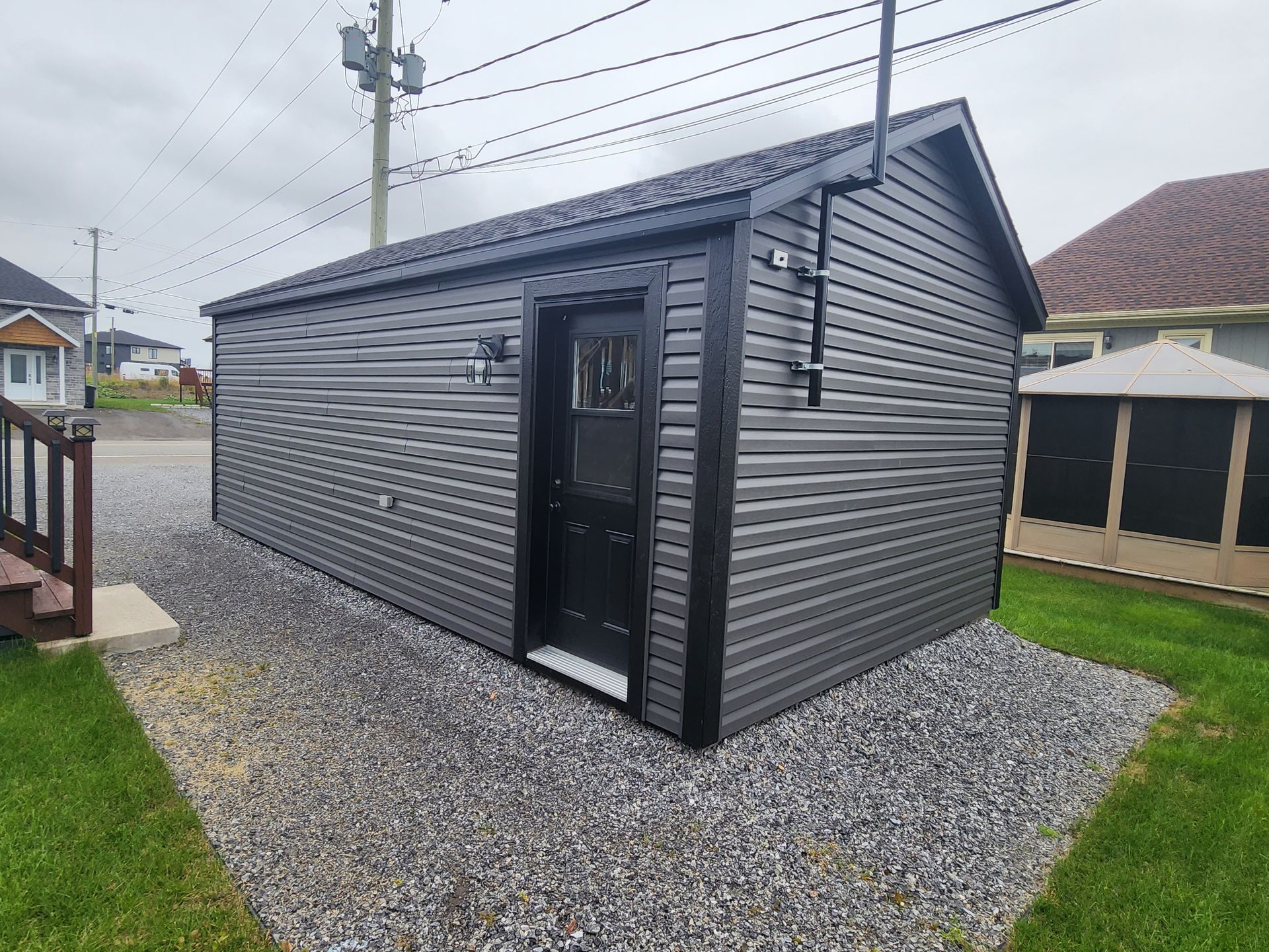 Un petit hangar avec une porte noire se trouve au sommet d'une allée de gravier à côté d'une maison.