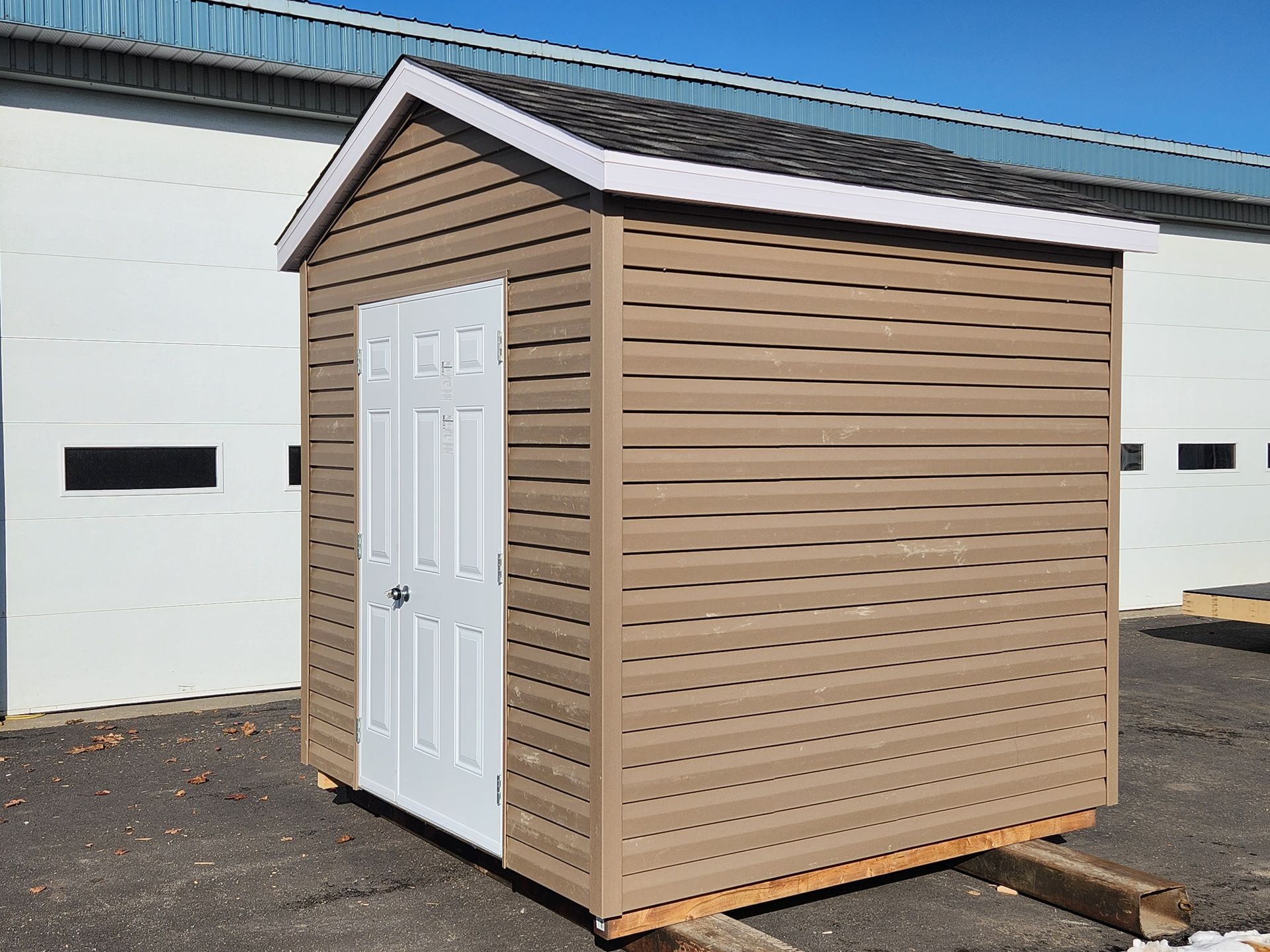 Un petit hangar marron avec une porte blanche se trouve devant une porte de garage blanche.