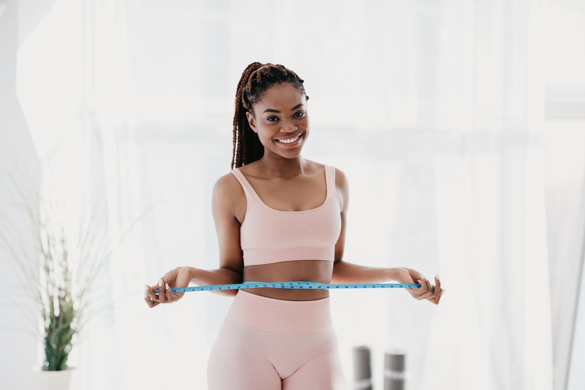 Woman in pink athletic wear smiles, measuring her waist with a blue tape measure.