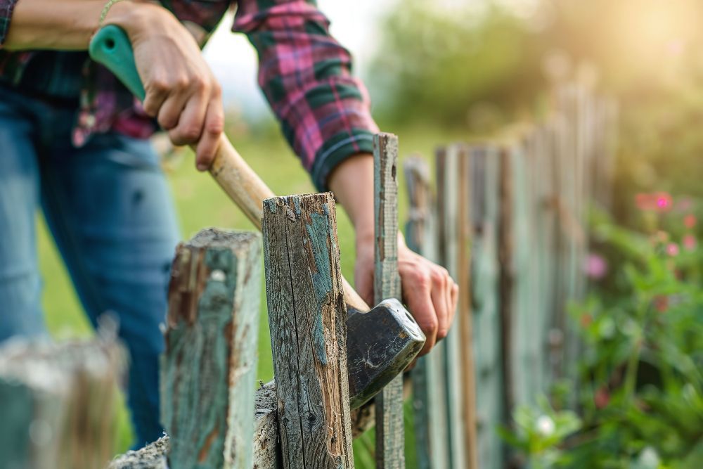A person wearing a plaid shirt uses an axe to repair a weathered wooden fence in a garden.