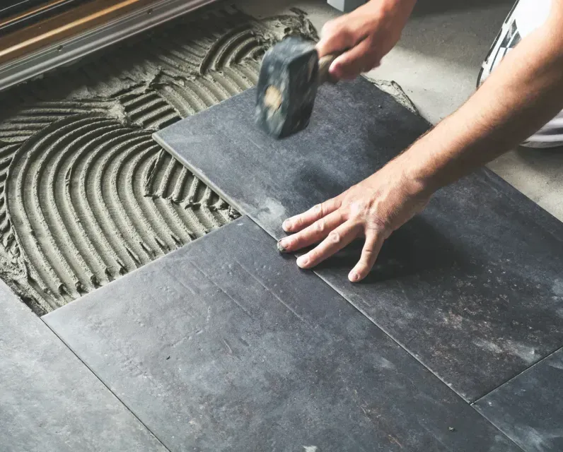 Hands laying dark floor tiles with a mallet over adhesive on the subfloor.