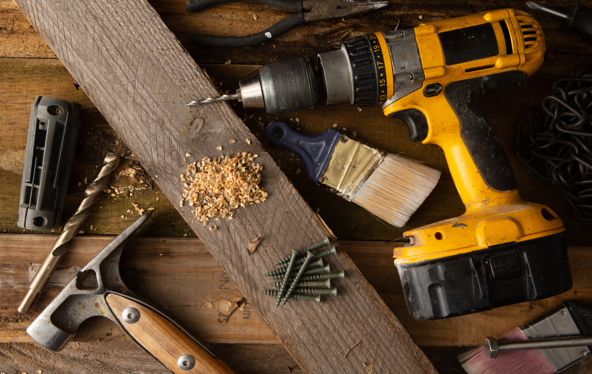 Yellow cordless drill, wood plank, screws, paintbrushes, and hand tools on a wooden workbench.